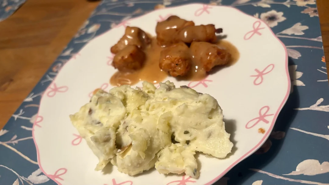 A plate with a serving of creamy mashed potatoes and several bacon-wrapped meatballs topped with brown gravy, placed on a blue floral placemat.