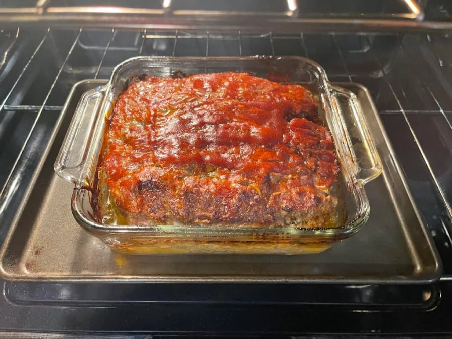 A glass baking dish with a cooked meatloaf topped with tomato sauce sits on a baking sheet inside an oven. The edges of the meatloaf appear slightly browned.