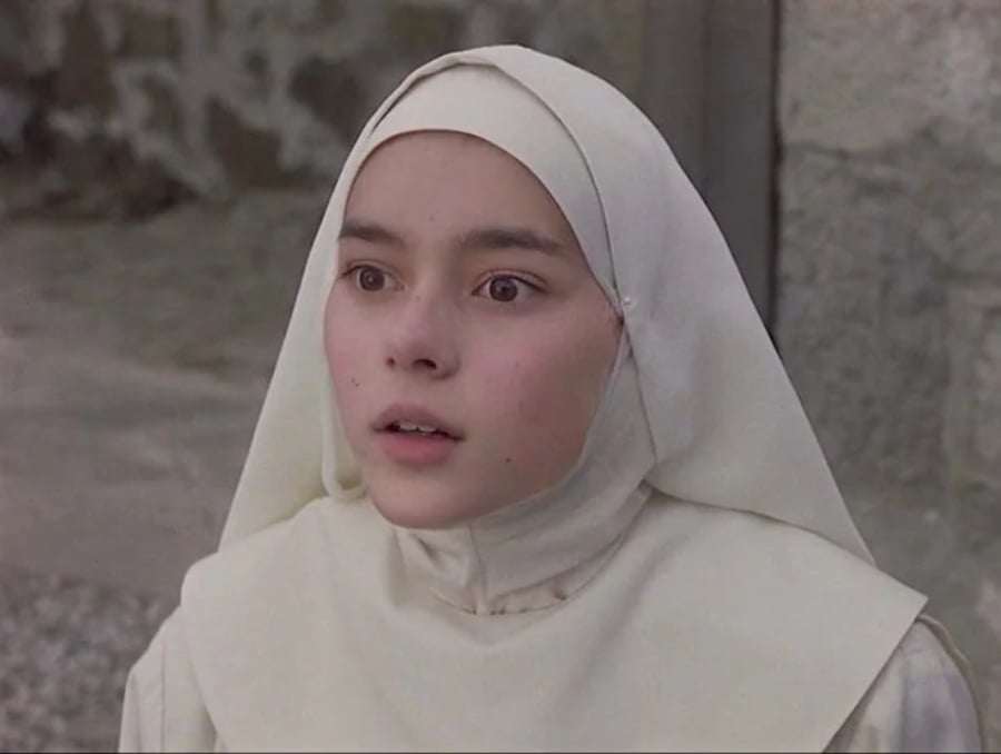 A young person dressed in a white nun’s habit and veil looks ahead with a surprised or amazed expression, standing outdoors near a stone wall.