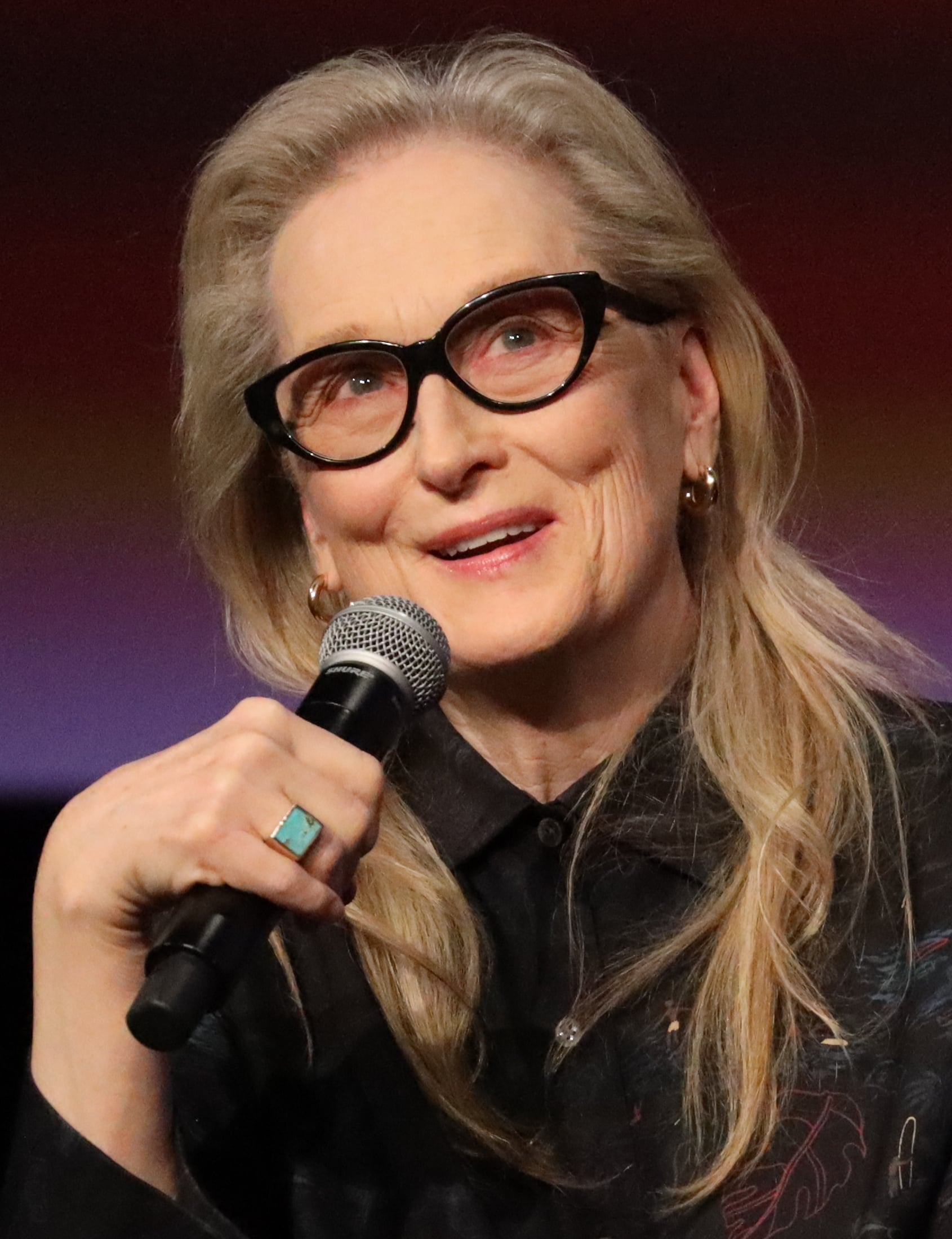 A woman with long blonde hair and black glasses speaks into a microphone. She is wearing a dark shirt and a turquoise ring, smiling gently with a colored background behind her.