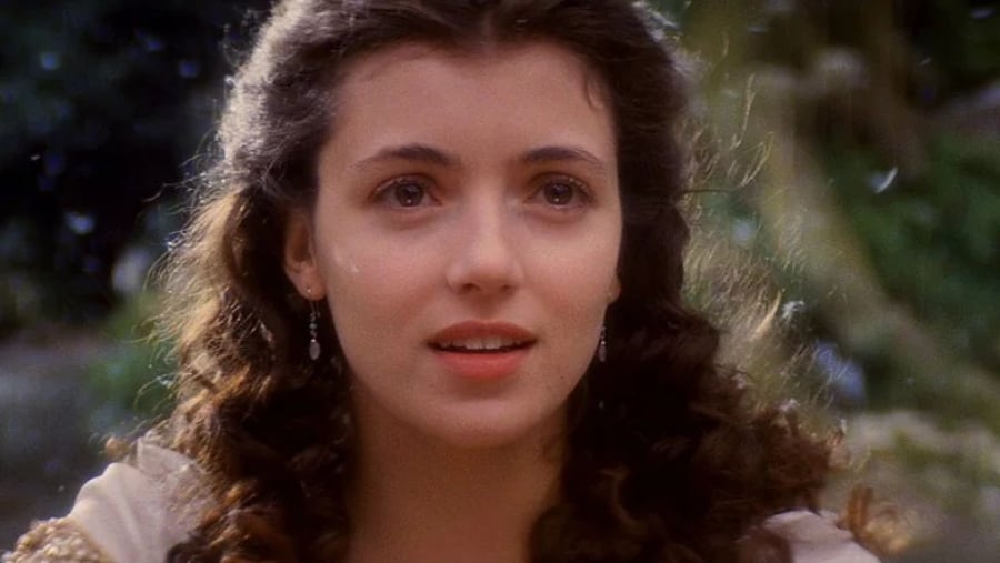 A young woman with long, curly brown hair and delicate earrings gazes softly ahead, surrounded by a natural, sunlit outdoor setting with greenery in the background.