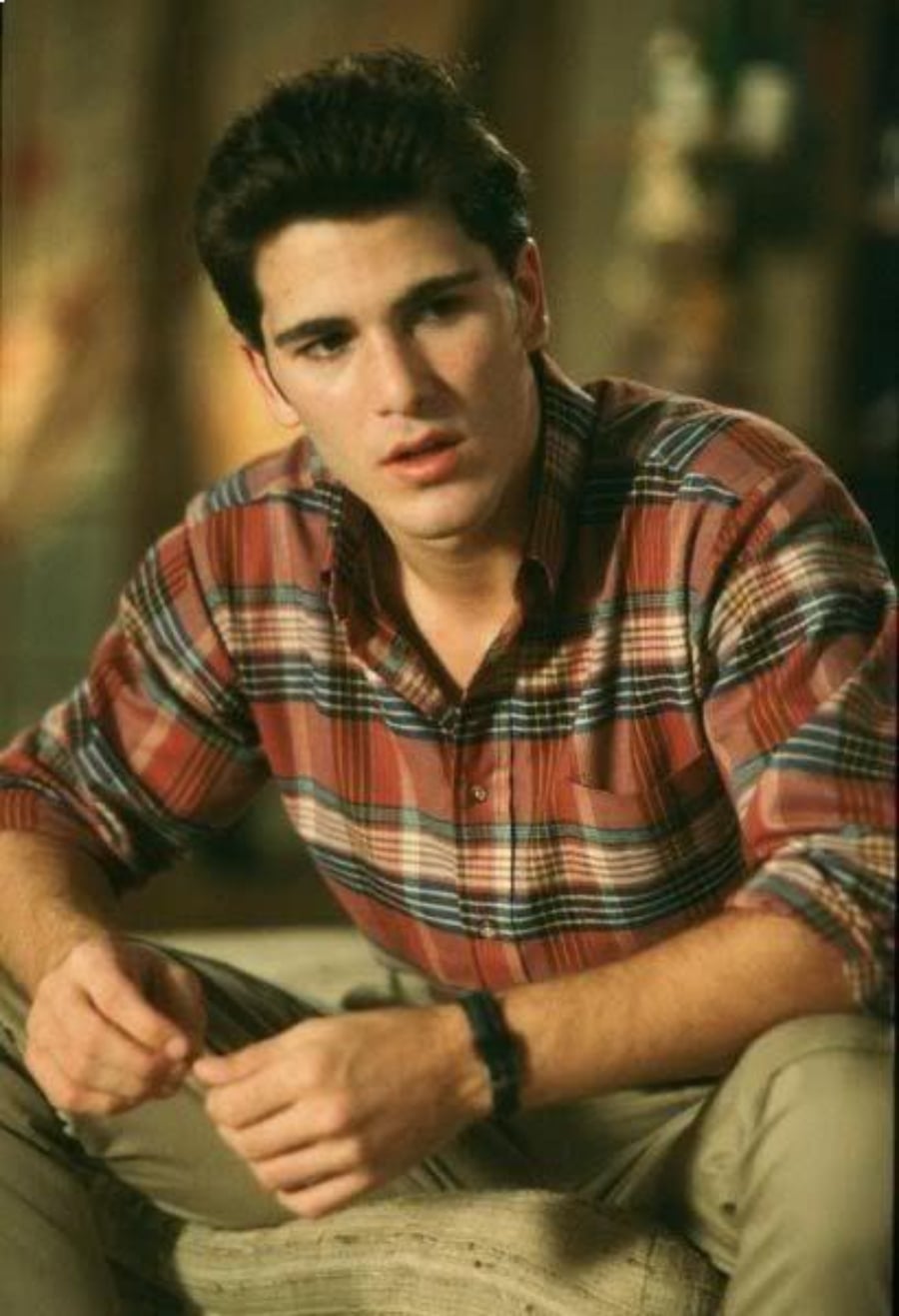 A young man with dark hair wearing a red plaid shirt sits indoors, looking slightly to the side with a thoughtful expression. His hands are resting in front of him, and the background is softly blurred.