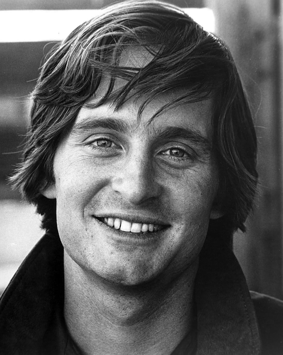 A young man with medium-length, tousled hair smiles at the camera in this black and white portrait. He is wearing a collared jacket and the background is softly blurred.