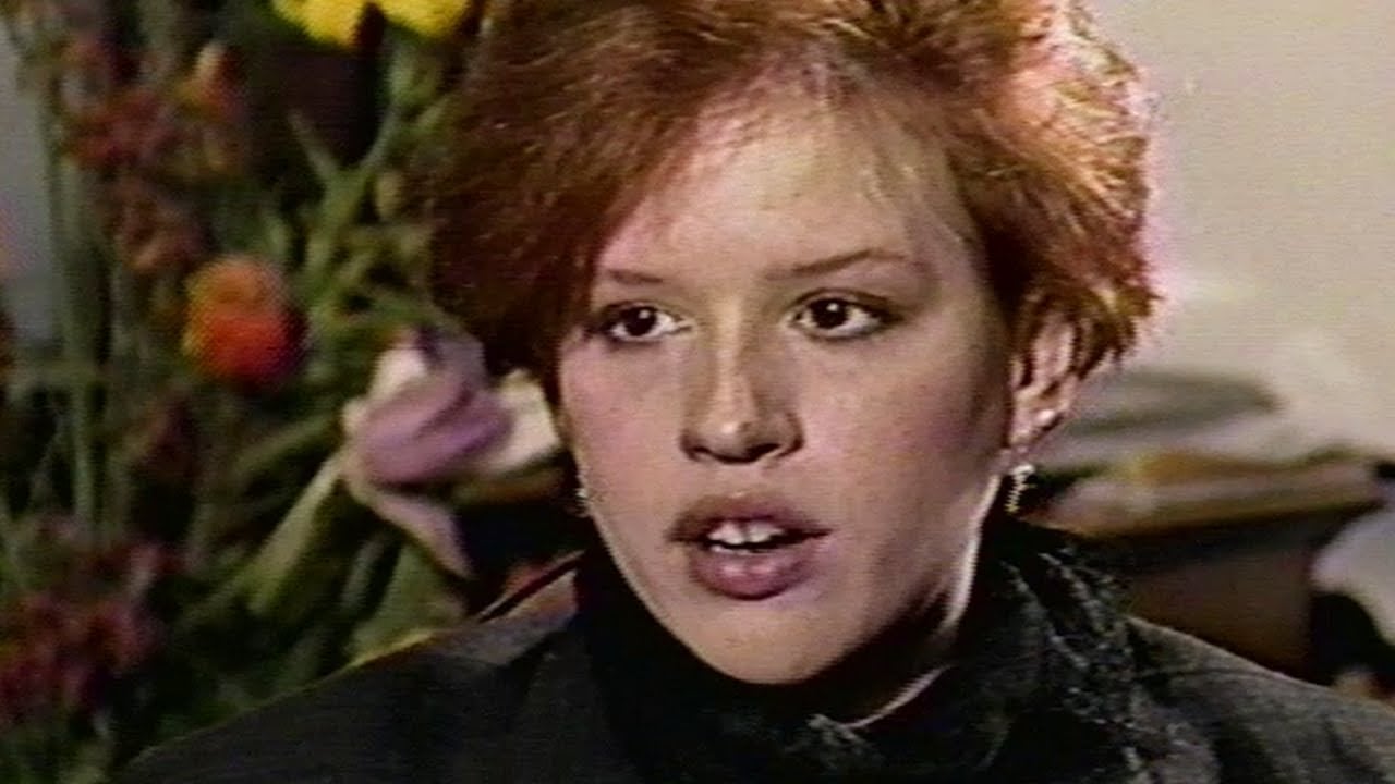 A person with short red hair and light skin is speaking, wearing a dark jacket and scarf. The background is blurred with colorful flowers visible. The lighting suggests an indoor setting.