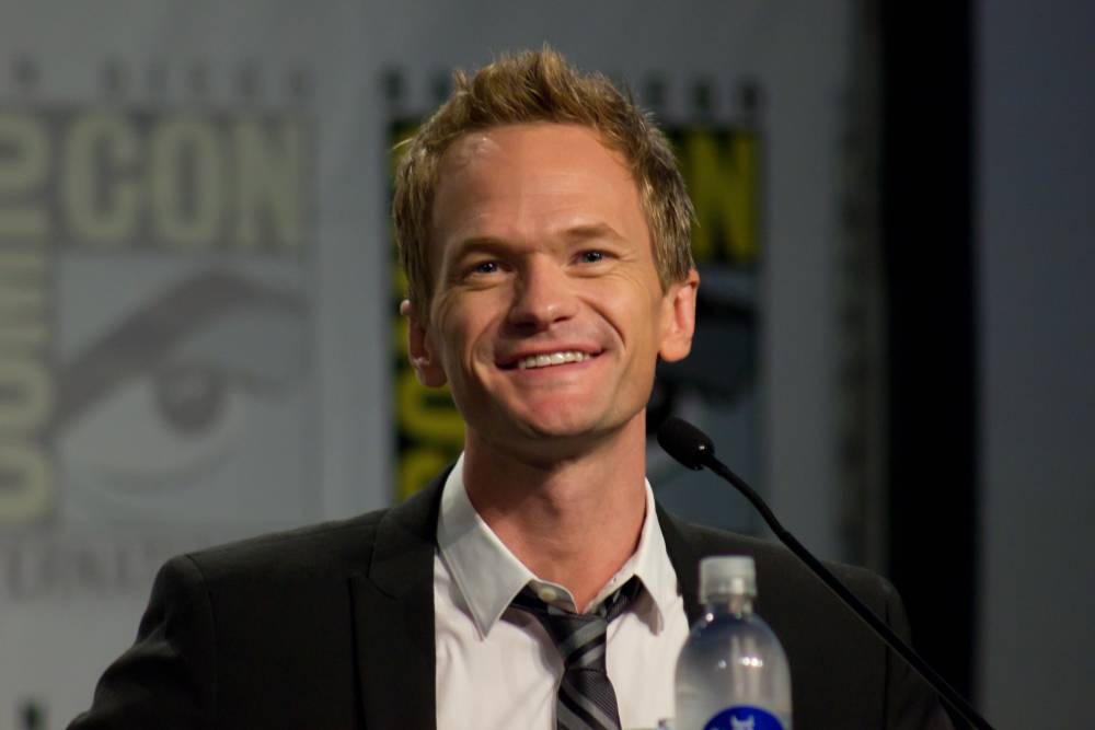 A man in a suit and tie smiles while speaking at a microphone. A water bottle is in the foreground, and a blurred Comic-Con logo is visible in the background.