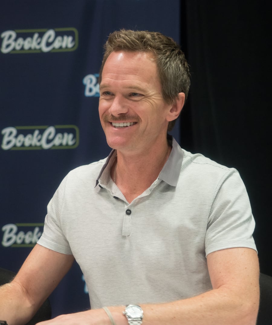 A smiling man with short brown hair in a light grey polo shirt sits at a table in front of a backdrop that reads "BookCon.