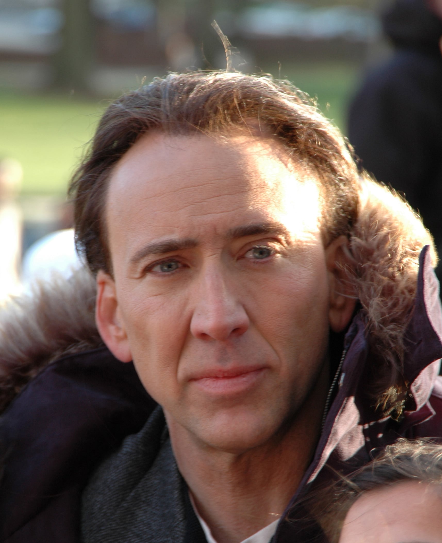 A man with light skin, brown hair, and blue eyes wears a dark coat with a fur-lined hood, standing outdoors in soft daylight.