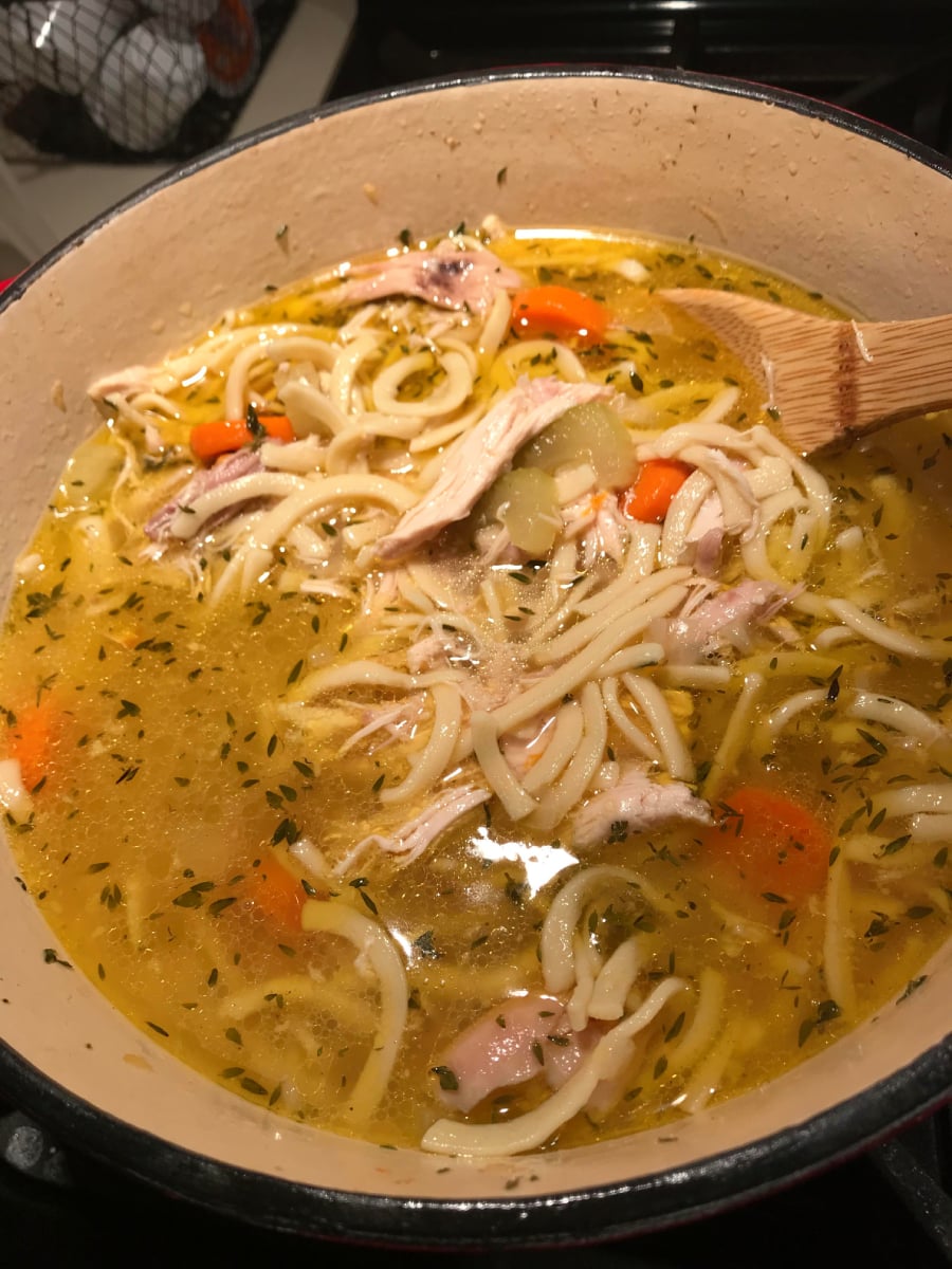 A pot of homemade chicken noodle soup with chunks of chicken, carrots, celery, and thick noodles in a seasoned broth, being stirred with a wooden spoon.