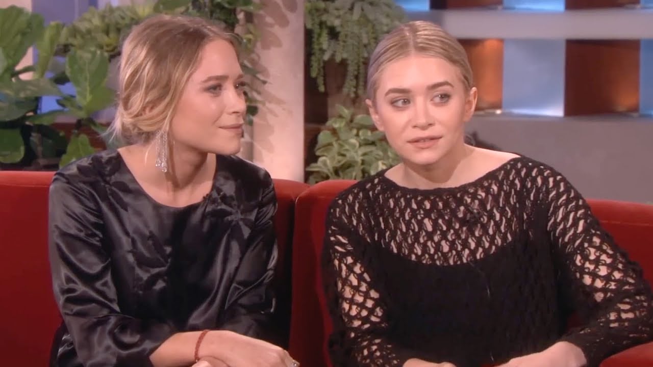 Two women with blonde hair sit next to each other on a red couch, engaged in conversation. One wears a black satin dress; the other wears a black knit top. Green plants and a softly lit studio background are visible behind them.