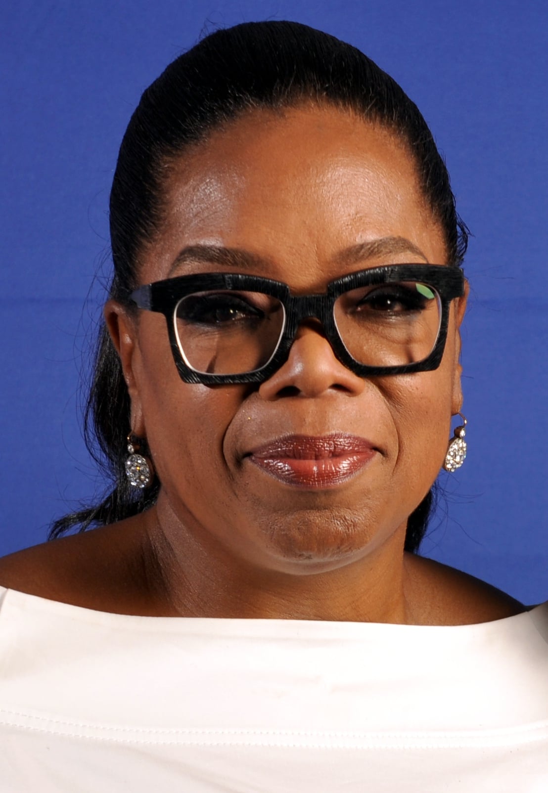 A woman with dark hair pulled back, wearing black glasses, diamond earrings, and a white top, stands in front of a blue background and smiles slightly.