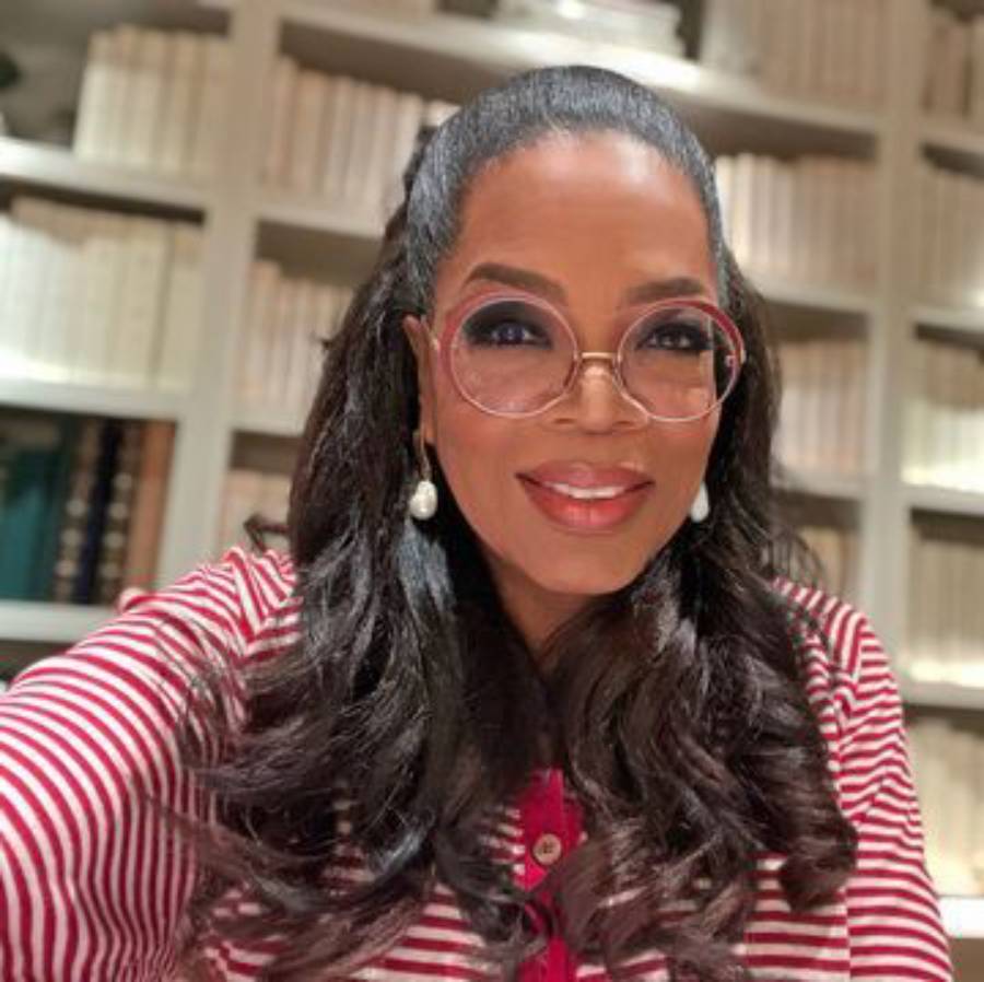 A woman with long dark hair, wearing round glasses, pearl earrings, and a red-and-white striped top, smiles in front of bookshelves filled with books.