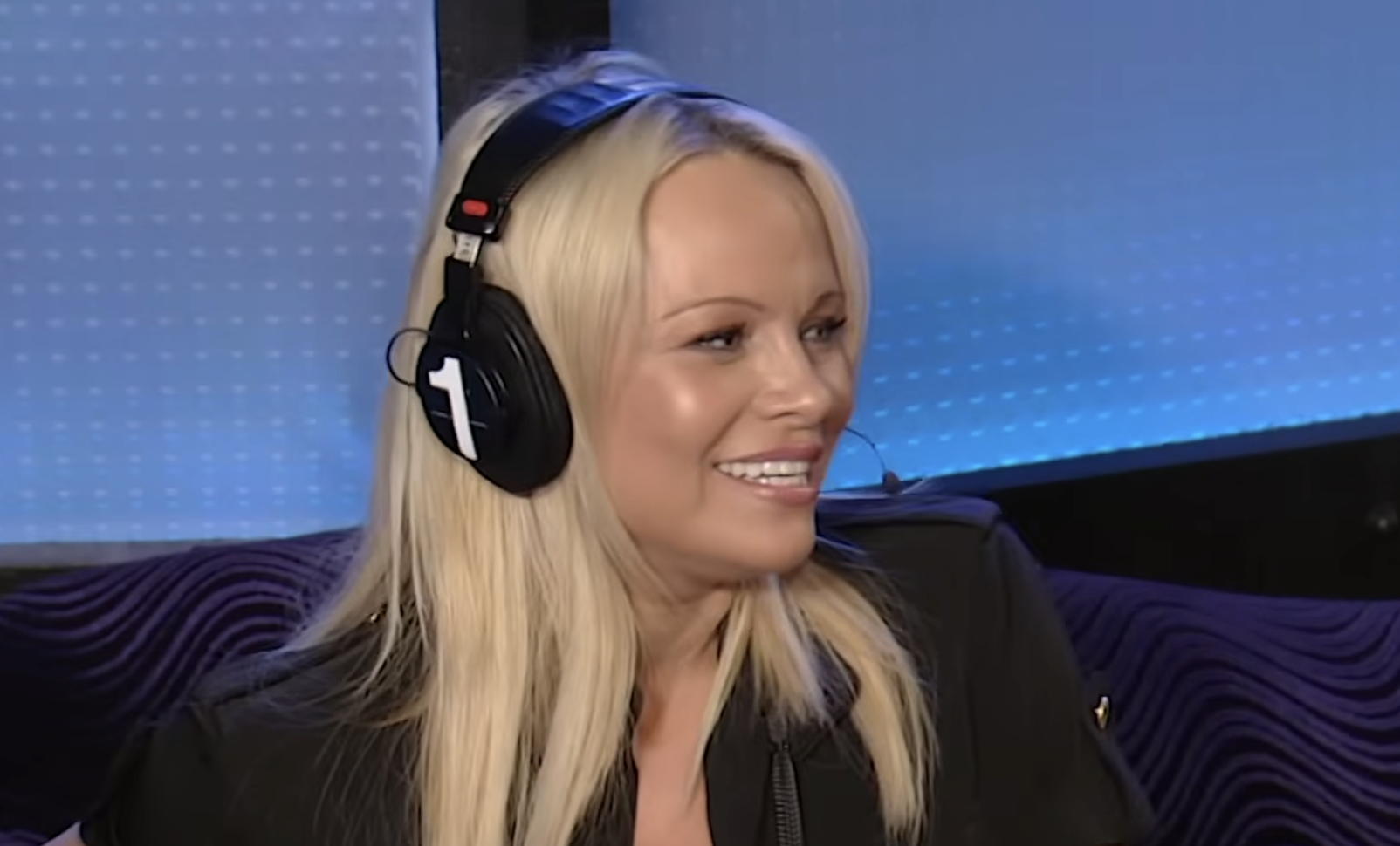 A woman with long blonde hair, wearing headphones with a microphone, smiles while sitting in a studio with blue lighting and a patterned wall behind her.