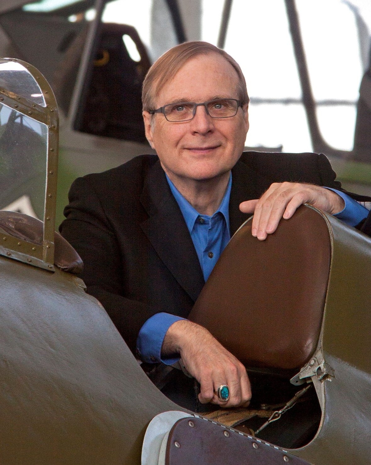 A man with glasses and a blue shirt under a dark blazer sits smiling in the cockpit of a vintage airplane, with one hand resting on the seat and the other visible with a turquoise ring.