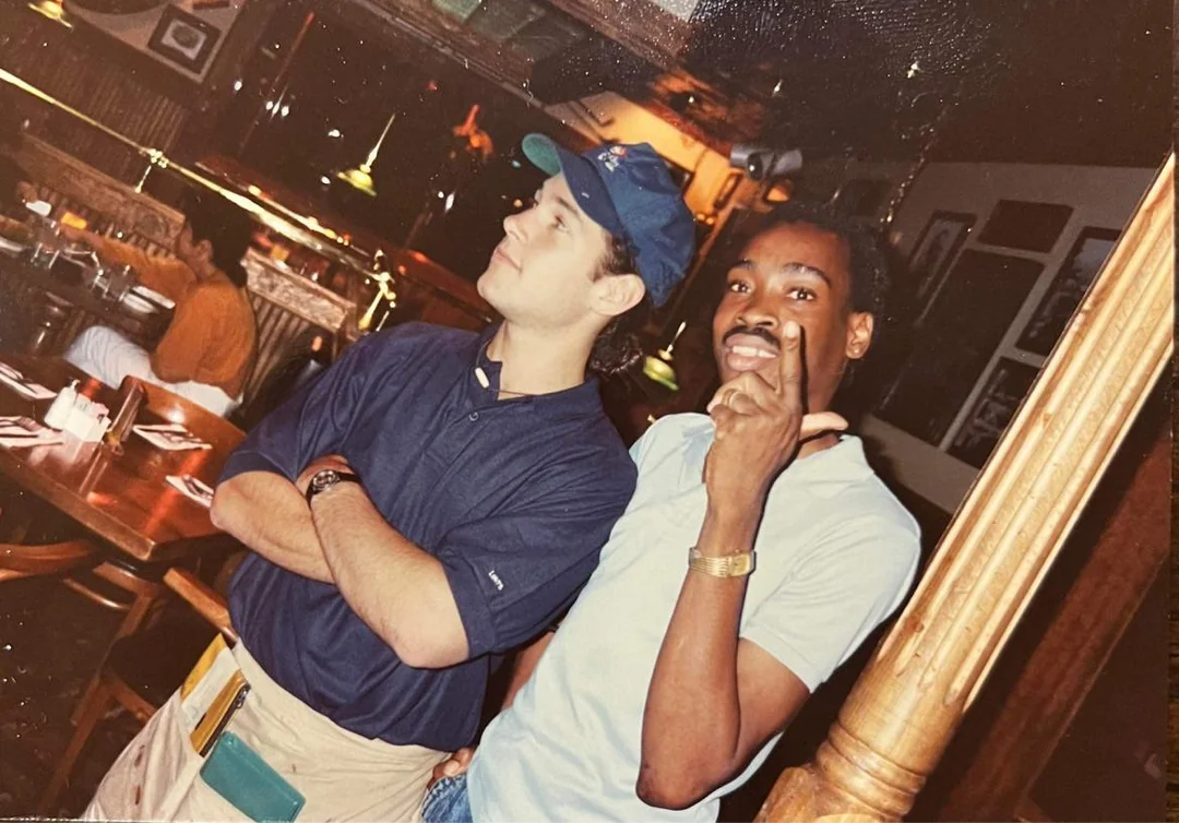 Two men pose inside a restaurant; one wears a blue shirt, cap, and apron, arms crossed, while the other, in a light blue shirt, smiles and makes a gesture with his hand. The restaurant has wooden furniture and dim lighting.
