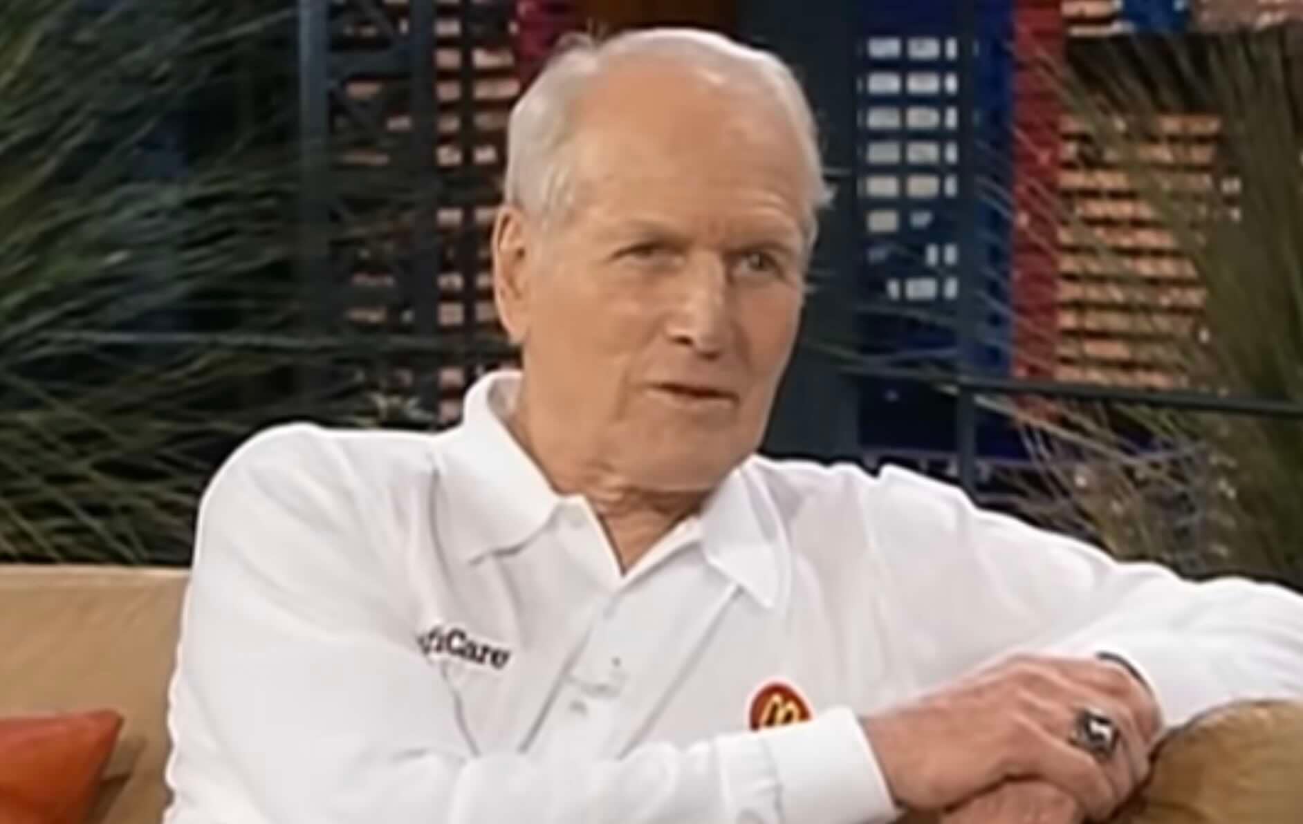 An elderly man with short white hair sits on a sofa, wearing a white collared shirt with embroidered logos. He is talking and gesturing with one hand, with a blurred background of indoor plants and decor behind him.