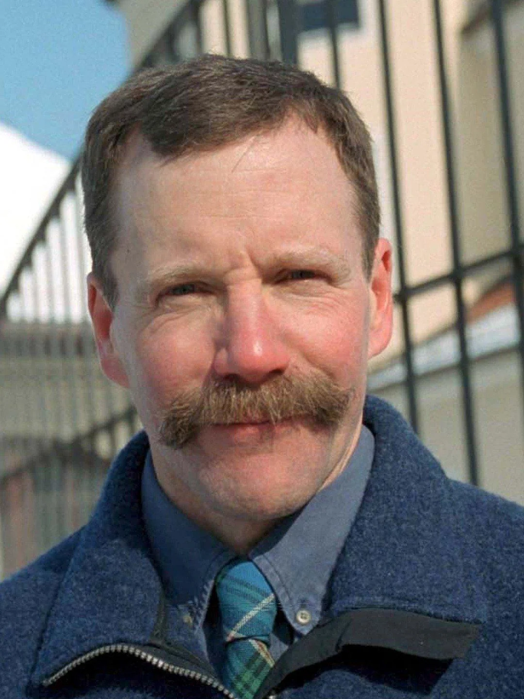 A man with a thick mustache and short brown hair stands outdoors, wearing a blue jacket and plaid tie. A black metal fence and buildings are blurred in the background under a clear sky.