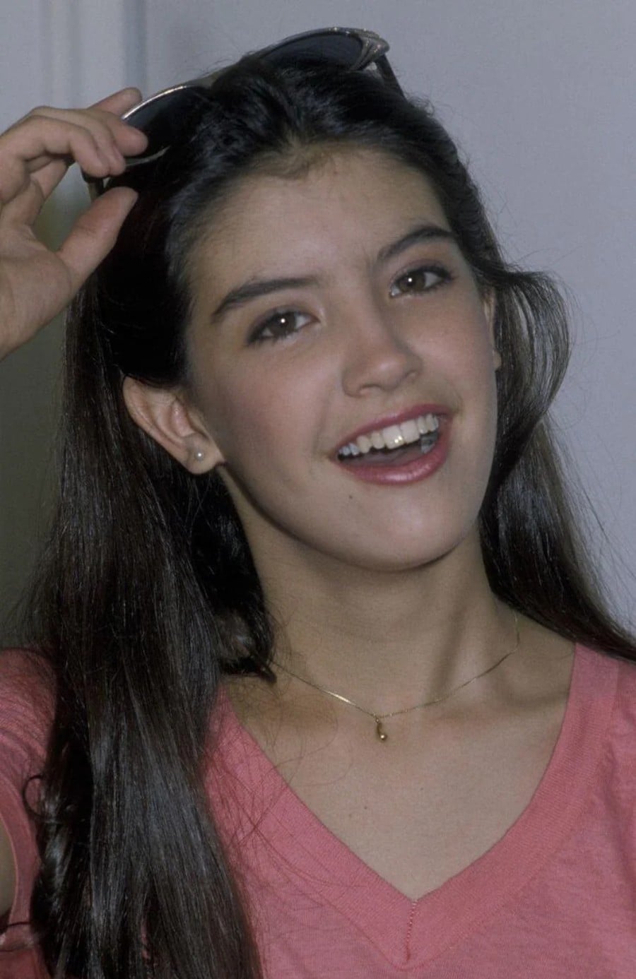 A young woman with long dark hair and a pink top smiles while holding sunglasses above her head. She is wearing a small necklace and appears relaxed and cheerful against a plain background.