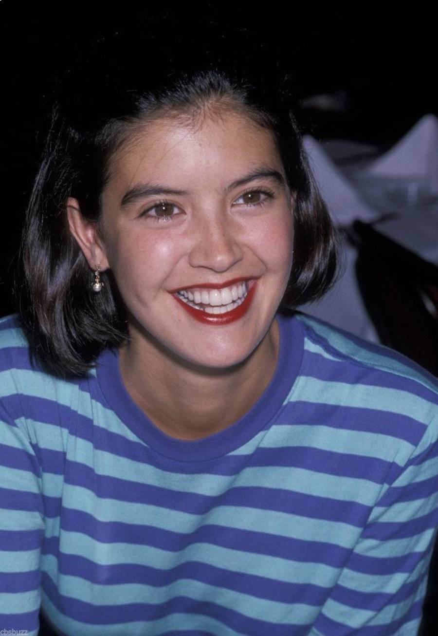 A young woman with short dark hair smiles brightly. She is wearing a blue and purple striped shirt and small hoop earrings. The background is dark and out of focus.