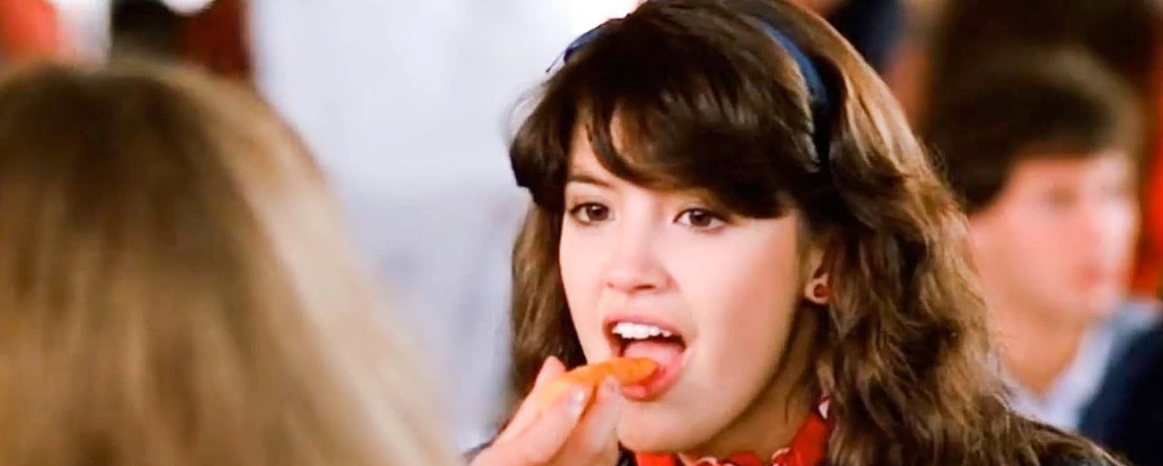 A young woman with dark hair and a blue headband is about to eat a carrot stick that another person is holding out to her. She is in a crowded setting, possibly a cafeteria.