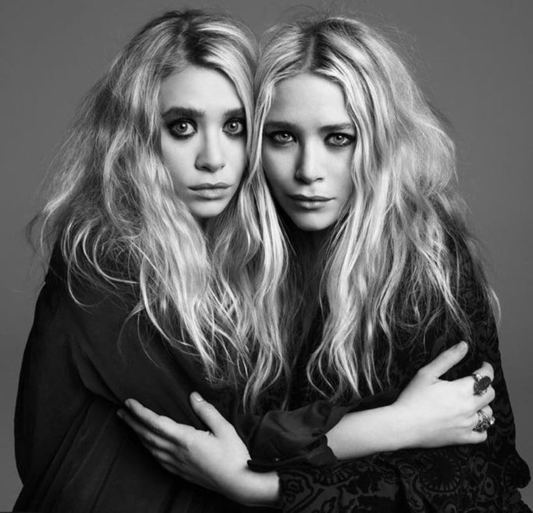 Two young women with long, wavy blonde hair and dark eye makeup pose closely together, wearing dark clothing and embracing each other in a black-and-white studio portrait.