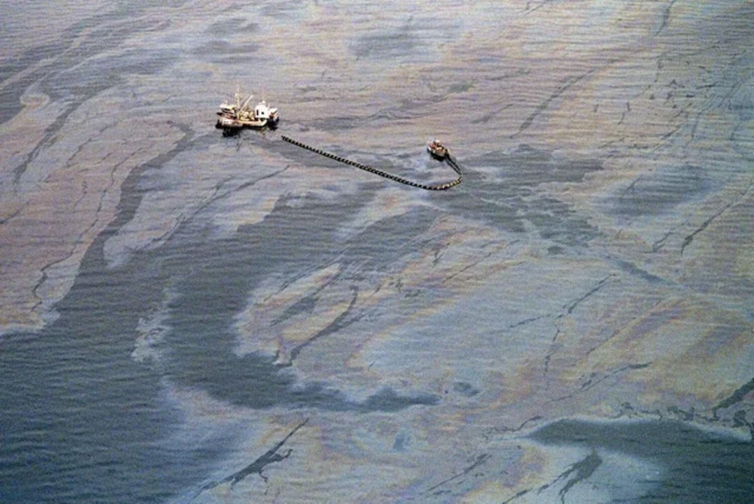 Aerial view of two boats in the ocean surrounded by large patches of oil slick on the water’s surface, indicating a significant oil spill. The boats appear to be involved in cleanup or containment efforts.