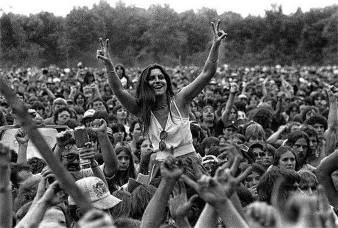 A large outdoor crowd cheers as a smiling woman, sitting on someone’s shoulders, raises both hands showing peace signs. The atmosphere is lively, and trees are visible in the background.