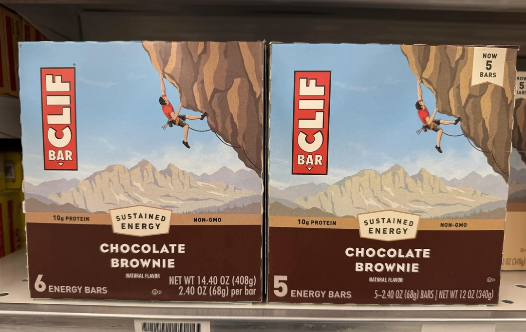 Two boxes of Clif Bar Chocolate Brownie energy bars on a store shelf. Each box features an illustration of a climber hanging from a cliff; one box contains 6 bars, the other contains 5 bars.