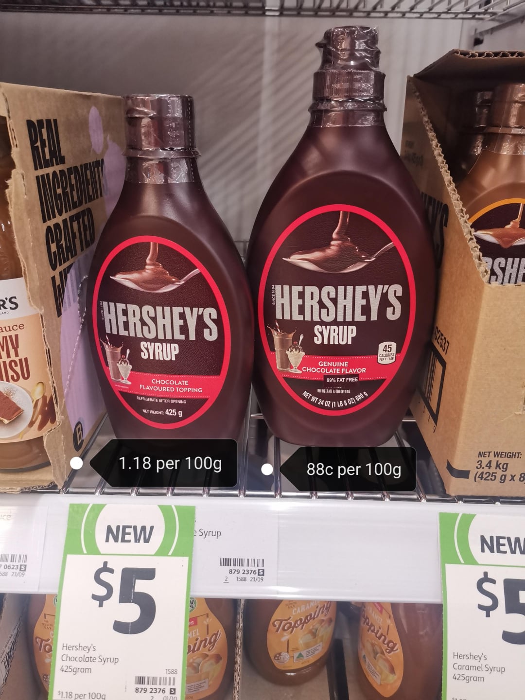 Two Hershey’s chocolate syrup bottles are on a supermarket shelf, each labeled $5. Price tags below show different prices per 100g: 1.18 AUD and 88c AUD. Other products are partially visible nearby.