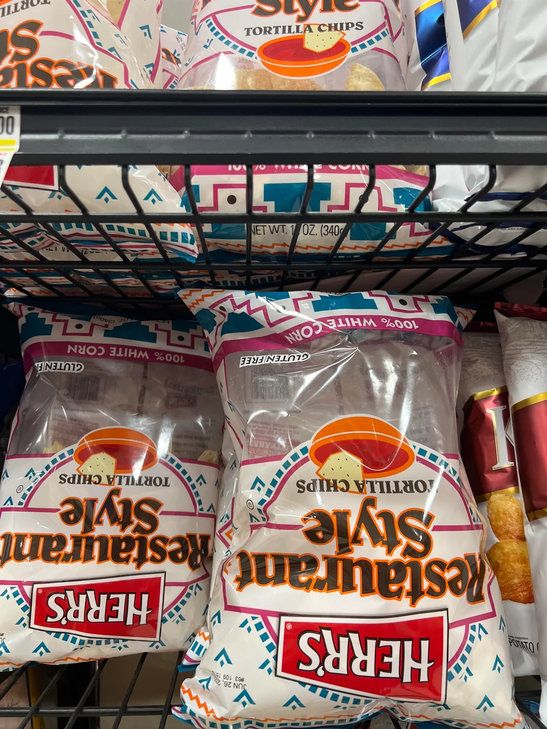Bags of Herr’s Restaurant Style Tortilla Chips are displayed on a grocery store shelf, with the red, white, and turquoise packaging visible. Some bags are upside down and next to other snack products.