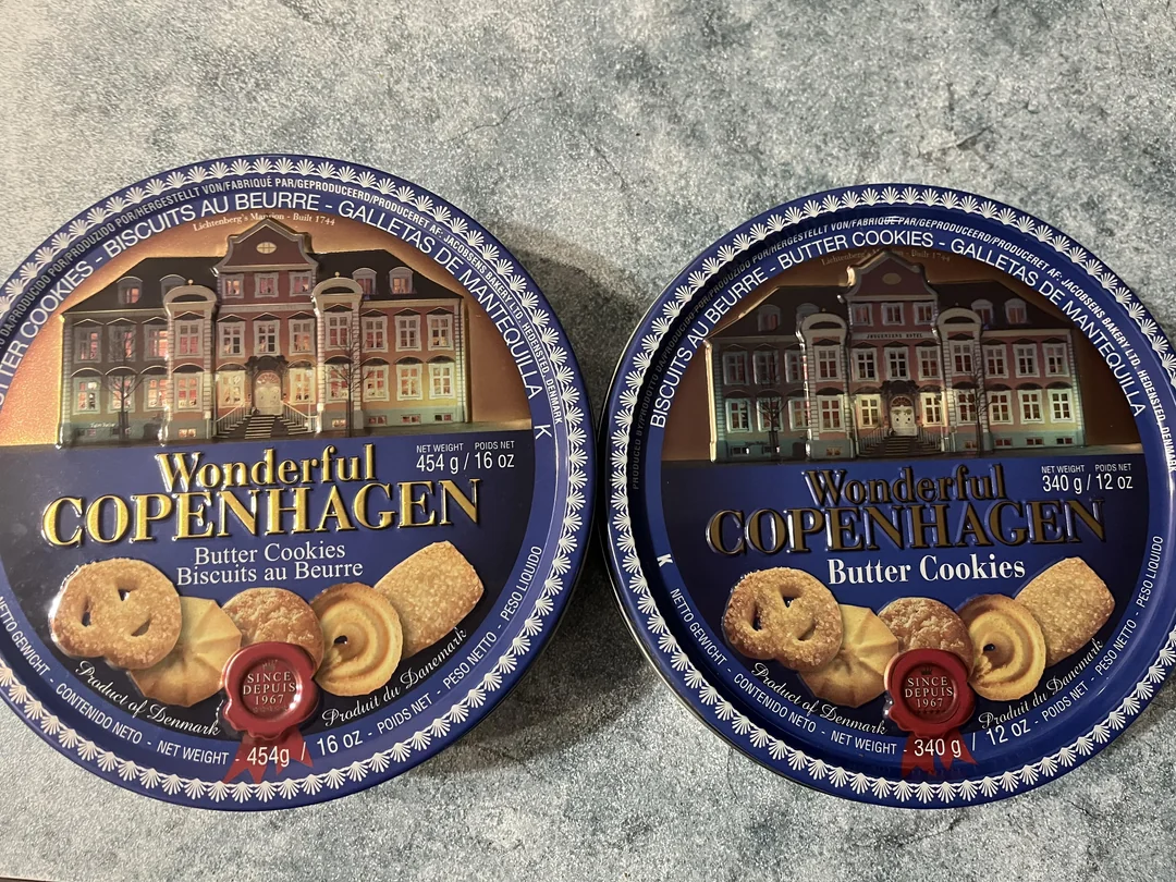 Two round blue tins of Wonderful Copenhagen butter cookies are shown on a gray countertop. The tins feature images of colorful European buildings and display their weights: 454g/16oz and 340g/12oz.