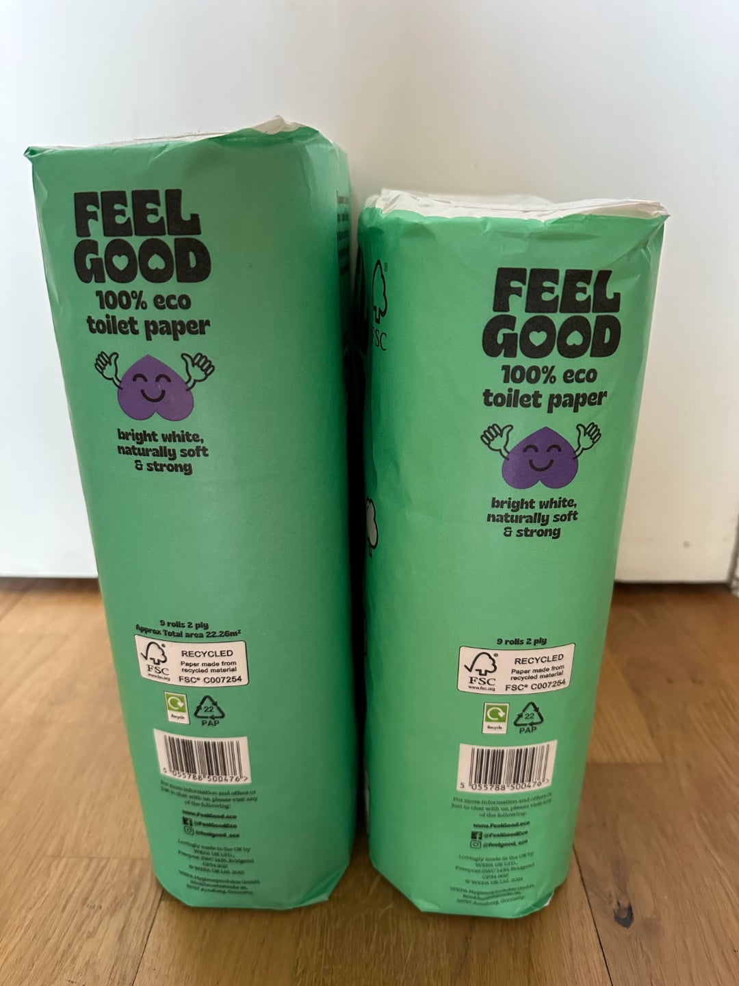 Two green packages of "Feel Good" 100% eco toilet paper stand side by side on a wooden floor. The packaging features a purple cartoon sheep and eco-friendly labeling.
