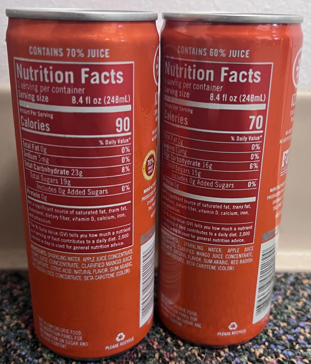 Two orange soda cans display nutrition facts labels side by side. The can on the left contains 70% juice and 90 calories per serving; the can on the right contains 60% juice and 70 calories per serving. Both cans are on a speckled countertop.