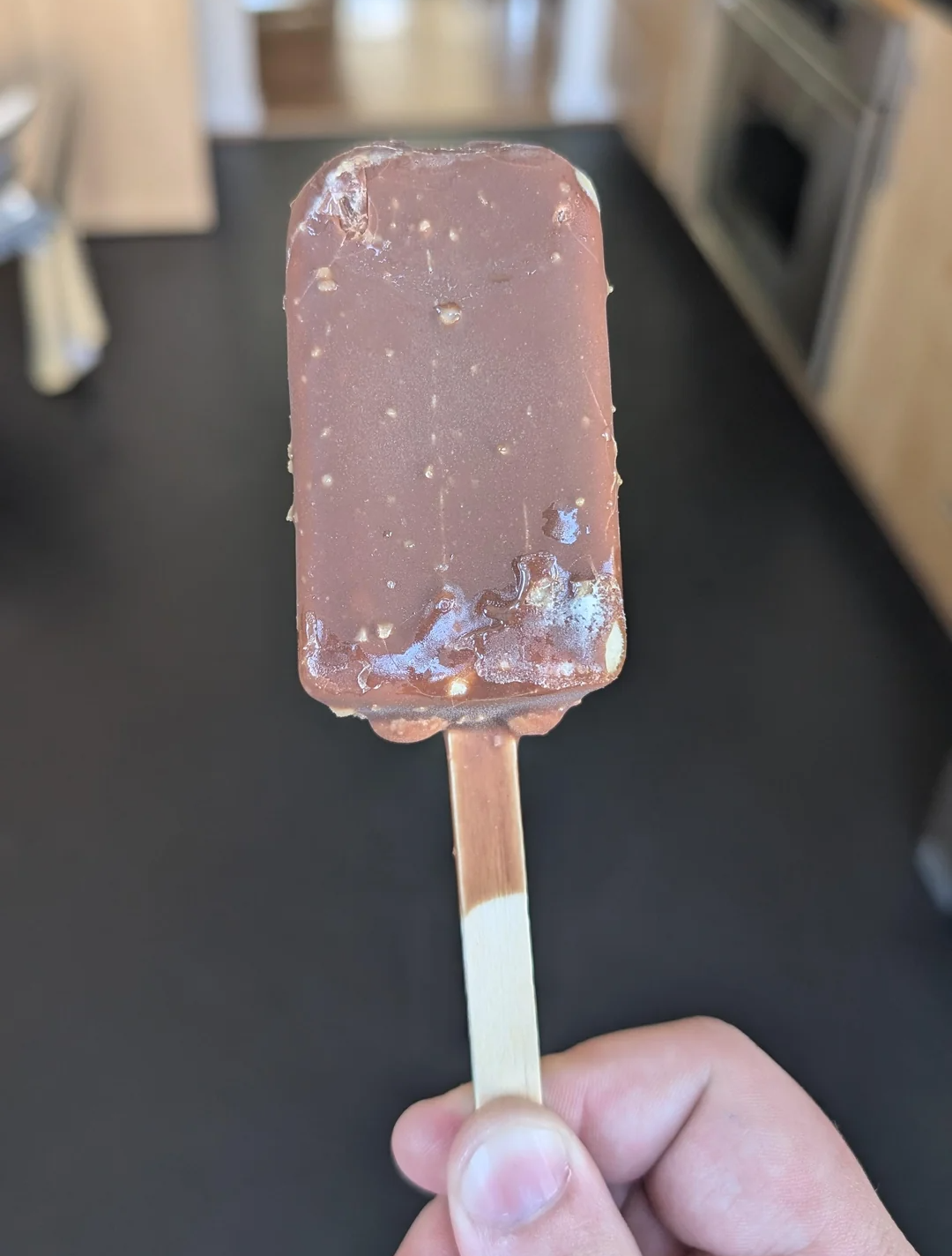 A hand holding a chocolate-covered ice cream bar on a wooden stick, with small bits visible in the chocolate coating. The background is a blurred kitchen scene.