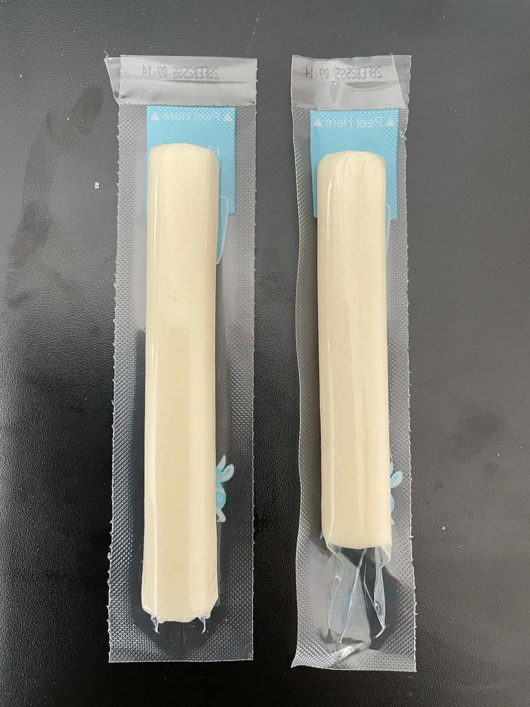 Two unopened sticks of string cheese in clear plastic wrappers are placed side by side on a dark surface. Both wrappers have a blue label on the top end.