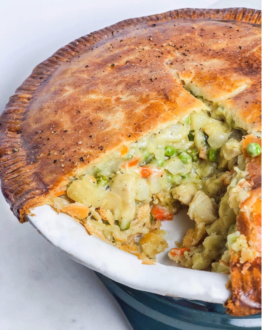 A golden-brown chicken pot pie with a slice removed, revealing a creamy filling of chicken, peas, carrots, and potatoes inside a flaky crust, displayed in a ceramic pie dish.