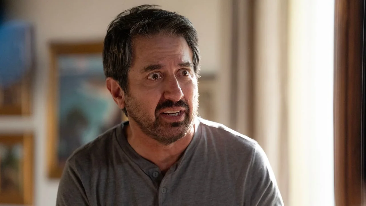 A middle-aged man with dark hair and a beard, wearing a gray henley shirt, looks concerned or surprised while indoors. Framed artwork and a soft curtain are visible in the background.