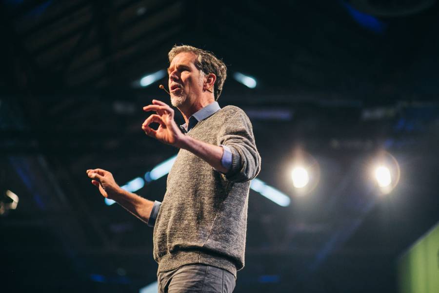 A man wearing a gray sweater gestures with his hands while speaking on a stage, with bright lights shining behind him and a dark, industrial-style ceiling overhead.
