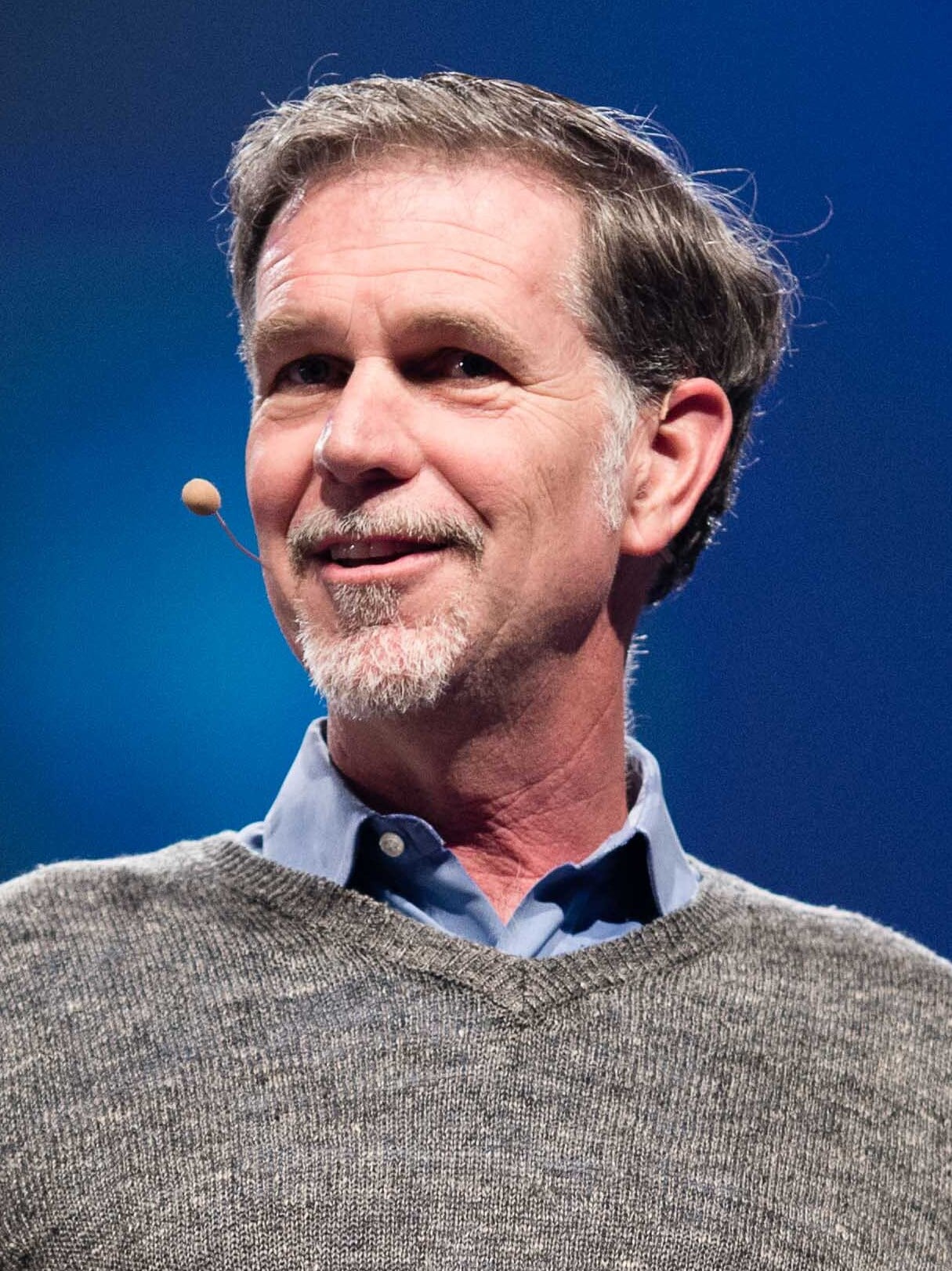 A man with short brown hair, a gray beard, and mustache is smiling while wearing a gray sweater over a blue collared shirt. He has a headset microphone and stands in front of a blue background.