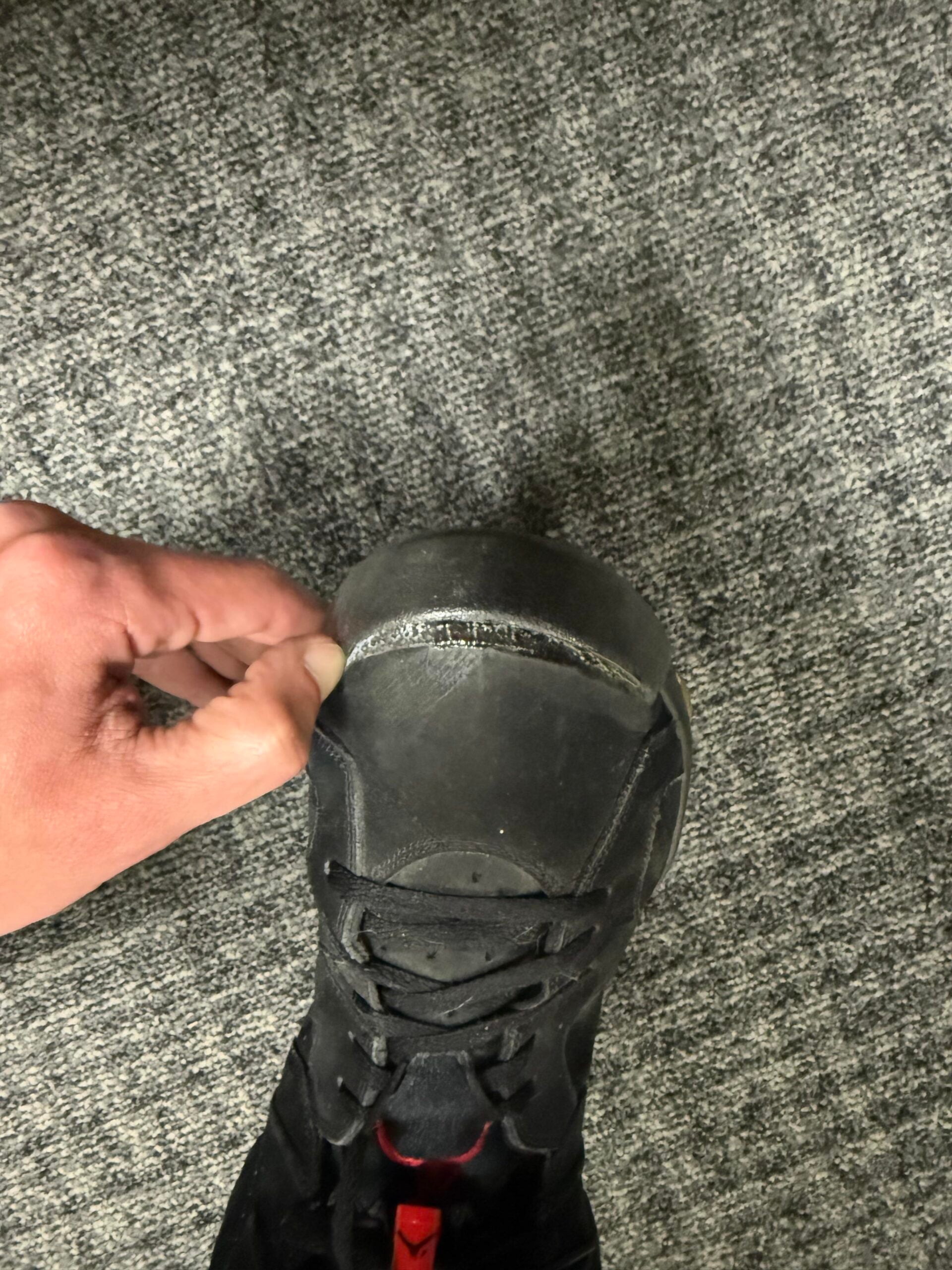 A hand lifts the toe of a black shoe, revealing a split seam or separation between the sole and upper part of the shoe, against a gray carpeted floor.
