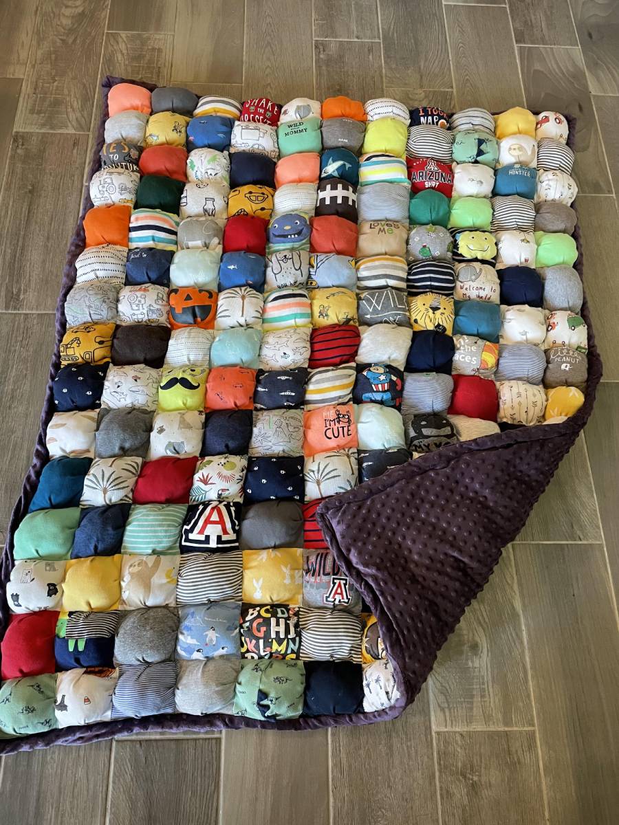 A colorful patchwork puff quilt made from various patterned and solid fabric squares, with a soft purple backing, is spread out on a tiled wooden floor. One corner of the quilt is folded over.