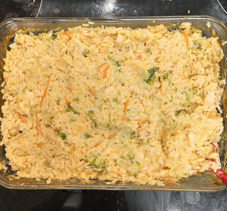 A glass baking dish filled with creamy rice casserole, mixed with shredded carrots, small broccoli pieces, and bits of chicken, all covered in a cheesy sauce.