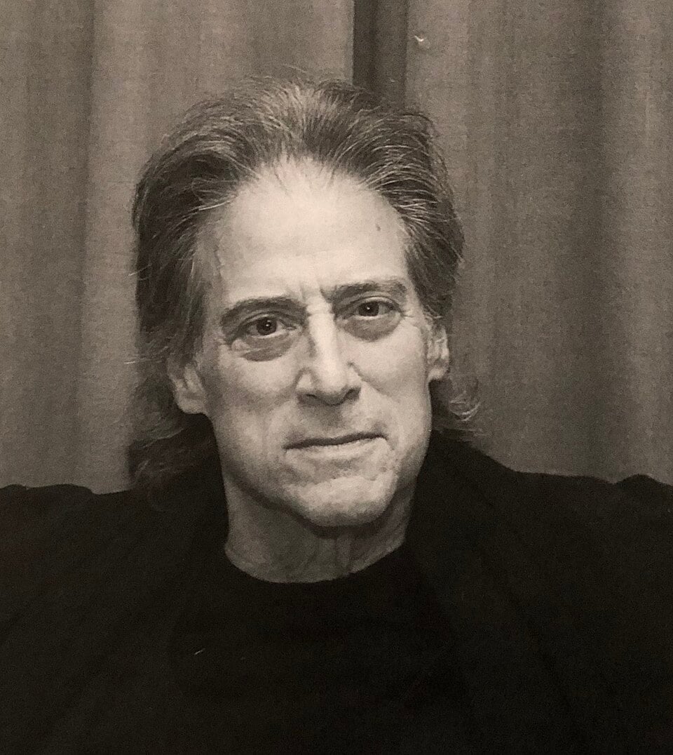 A black and white photo of an older man with gray hair, wearing a dark shirt, sitting in front of a curtain and looking directly at the camera with a neutral expression.