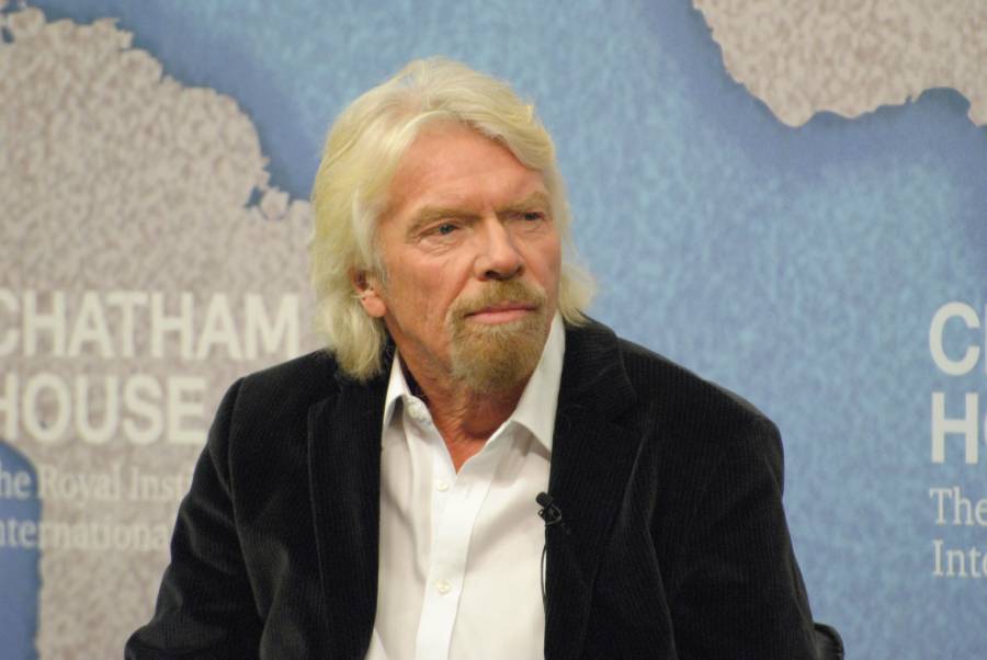 A man with white hair and a beard, wearing a white shirt and dark jacket, sits in front of a blue backdrop with the words "Chatham House" and a world map visible.