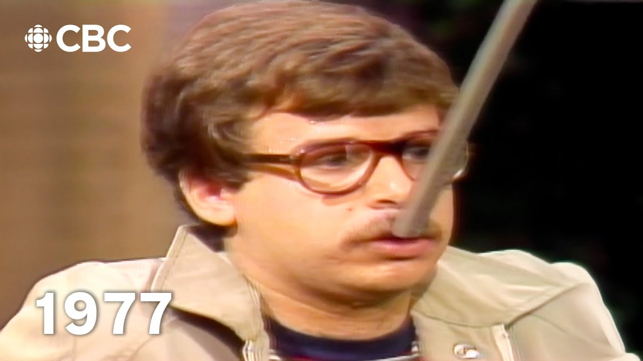 A man with brown hair, glasses, and a beige jacket appears with a long fake nose, reminiscent of Pinocchio, on a CBC television show. “1977” is displayed in white text at the bottom left.