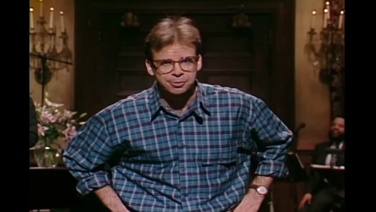 A man wearing glasses and a blue plaid shirt stands with his hands on his hips, looking forward. He is indoors, with dark wood paneling and plants visible in the background.