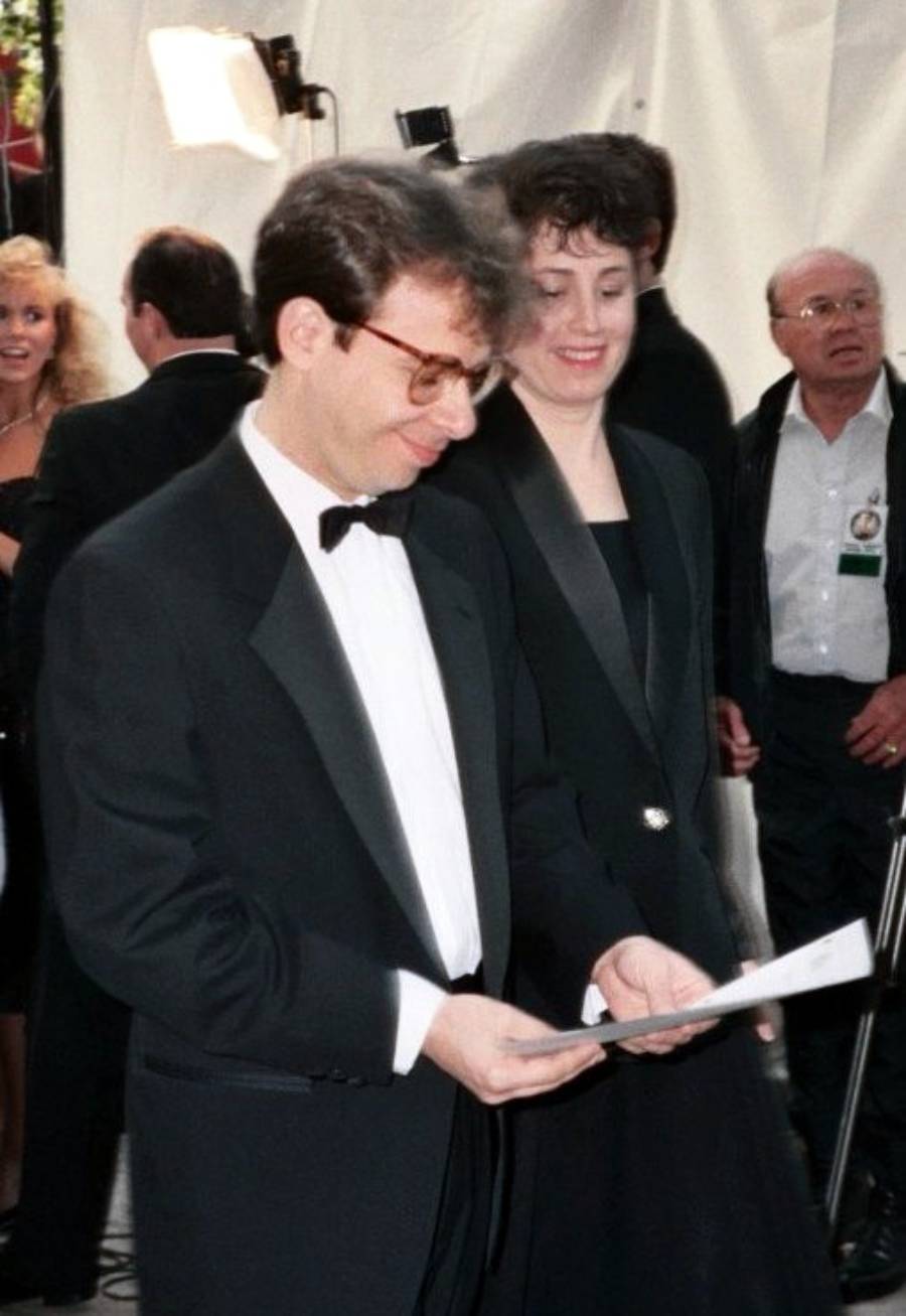 A man in a black tuxedo and glasses is holding papers and standing next to a woman in a black outfit at a formal event, with other people and photographers in the background.