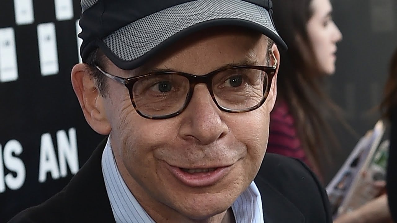 A man wearing glasses and a gray cap smiles while looking slightly to the side. He is dressed in a collared shirt and blazer. There are people and a dark event backdrop in the background.