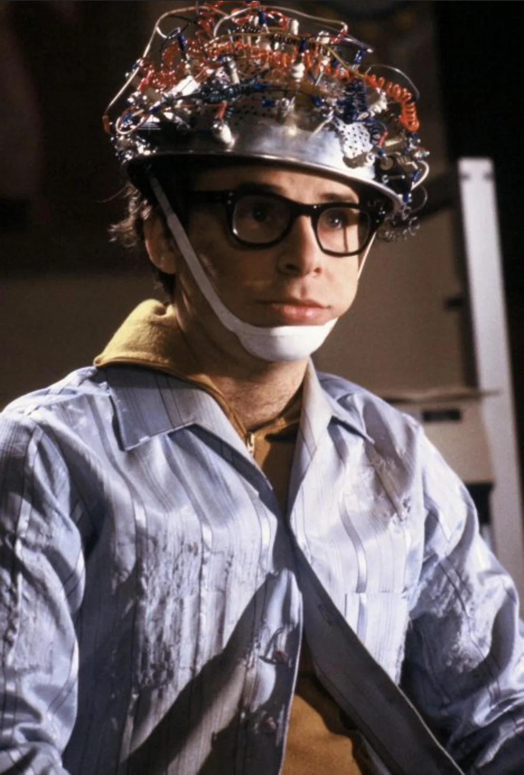 A person wearing large glasses and a headpiece made of wires and electronics sits indoors, looking forward with a neutral expression. They are dressed in a striped shirt and a chin strap secures the device to their head.