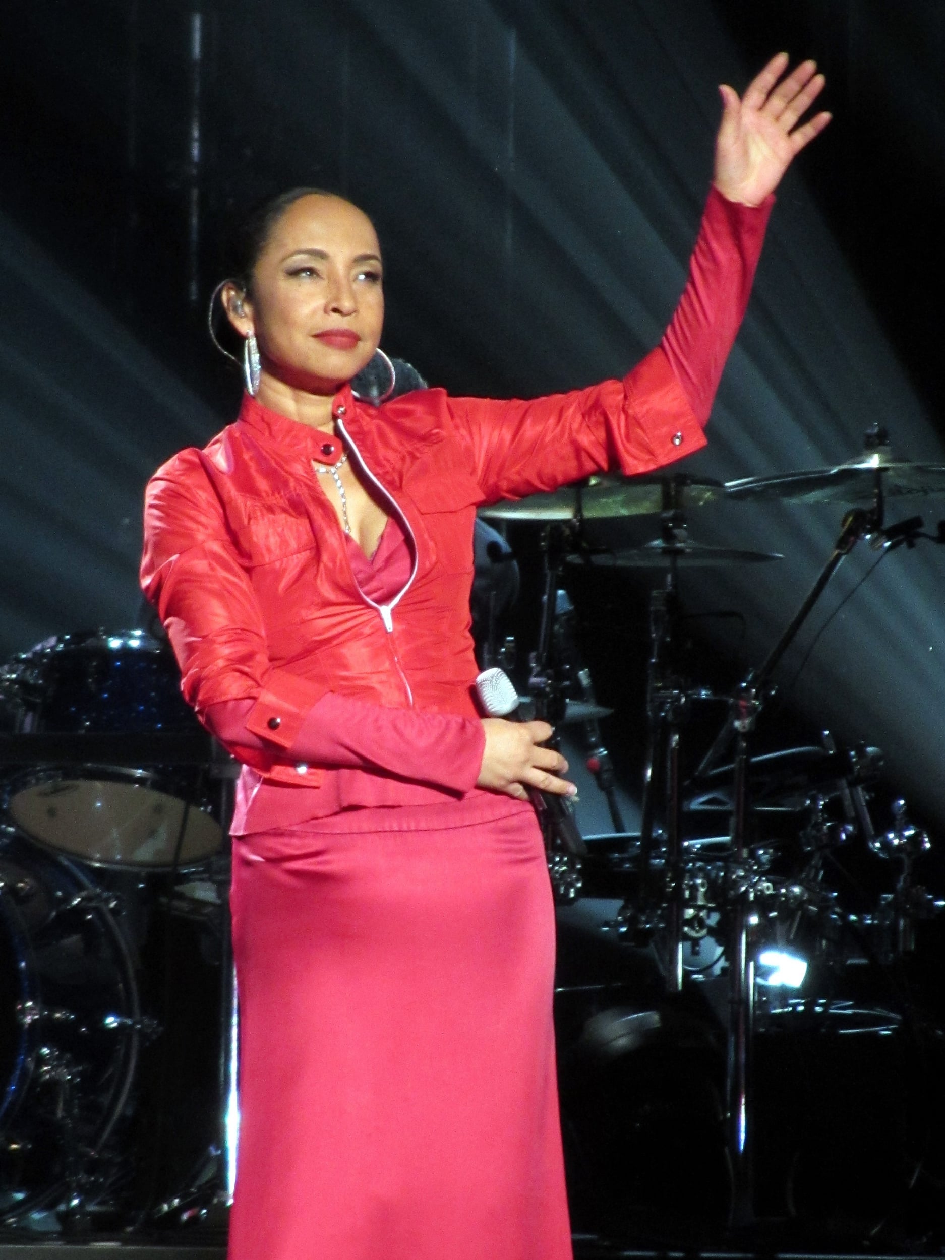 A woman wearing a bright red outfit stands on stage, holding a microphone in one hand and raising her other hand, with stage lights and drums visible in the background.