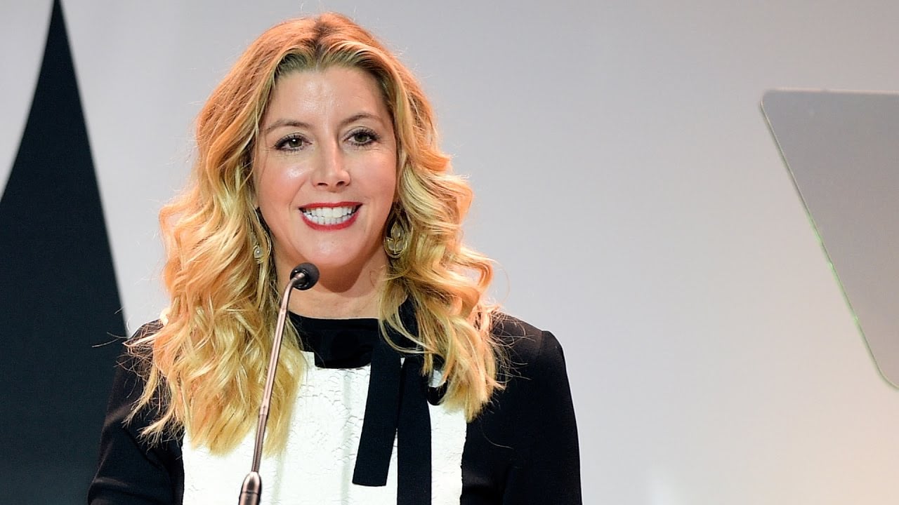 A woman with long, wavy blonde hair and a black-and-white outfit stands at a podium, smiling while speaking into a microphone against a light background.